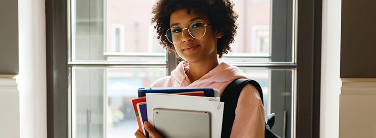 Student with glasses and backpack holding notebooks and tablet at school doors