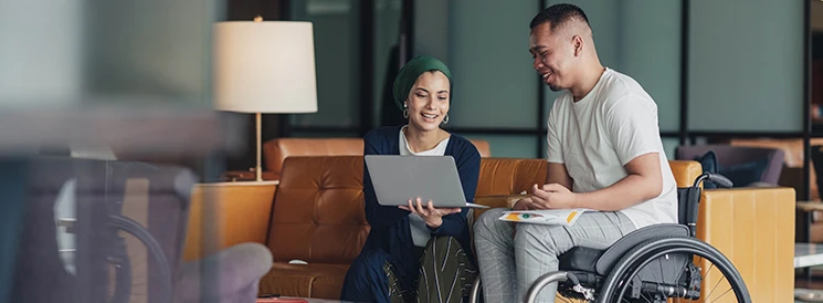 Man in a wheel chair working on a laptop with a woman on a couch