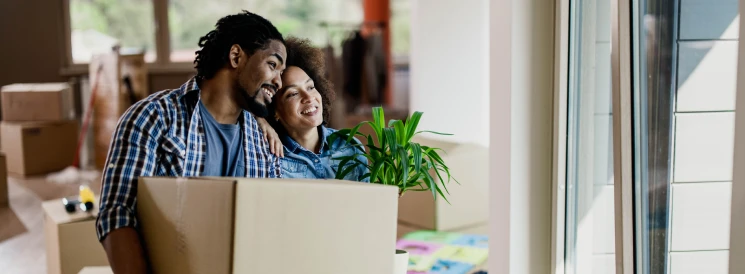 couple looking out window of new home holding moving box