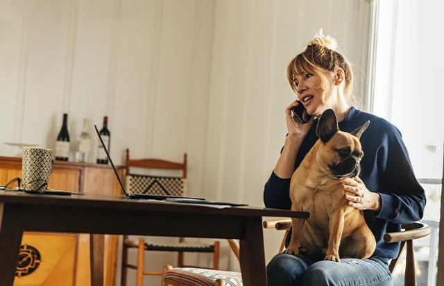 Woman sitting at kitchen table talking on phone with dog in lap