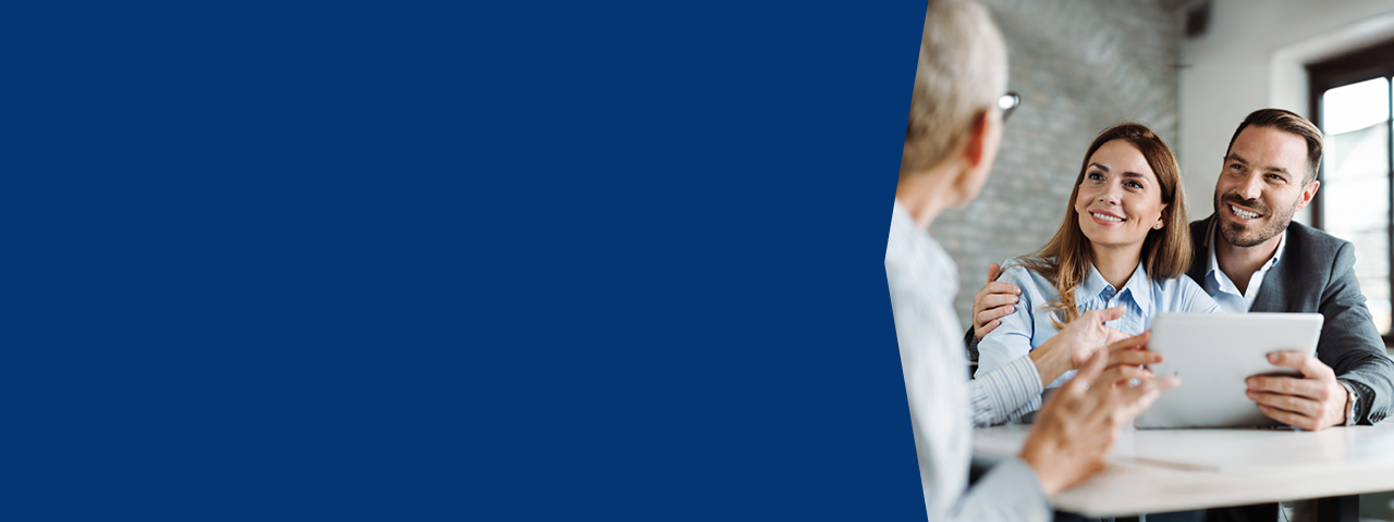 Image showing a professional meeting in a modern office setting. Two individuals sit side by side, holding a digital tablet, while another person across the table gestures as if explaining details. The scene suggests a financial planning or advisory discussion, possibly reviewing investment options or wealth management strategies. The right side of the image features the meeting participants, while the left half is a solid dark blue background, likely reserved for promotional text or branding.