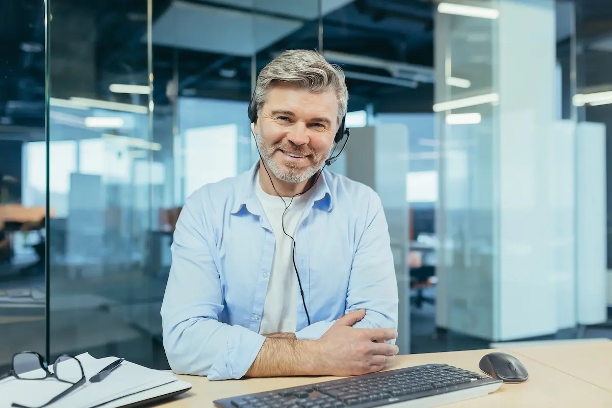Smiling customer support representative wearing a headset, seated at a desk in a modern office environment.
