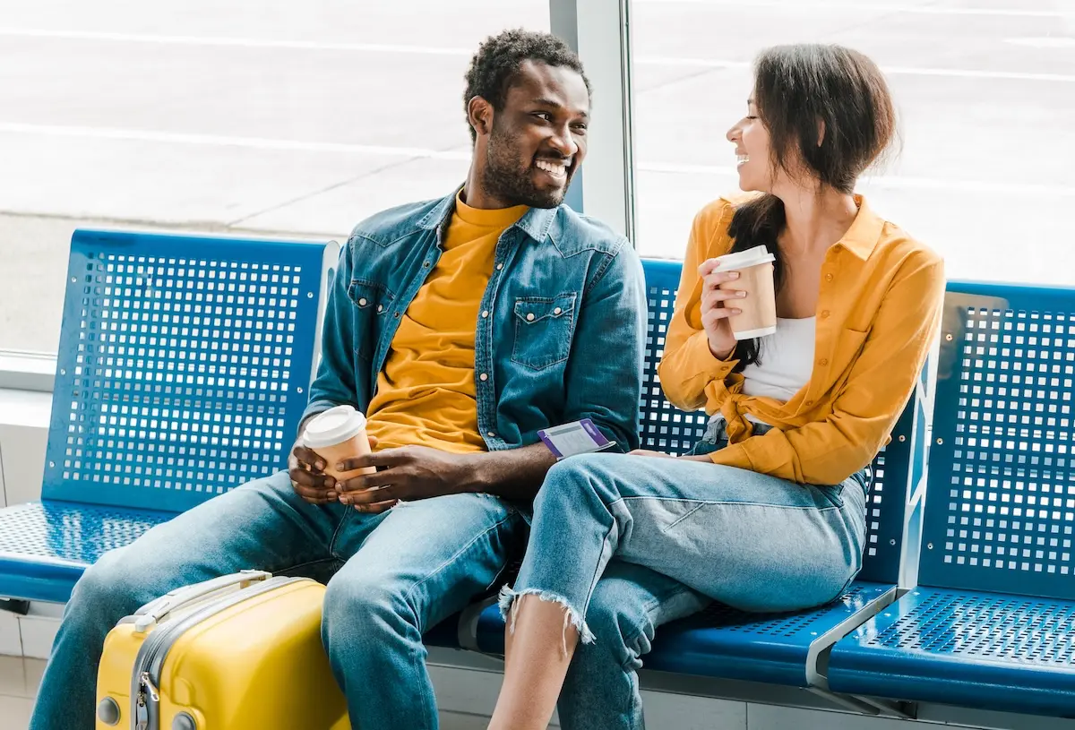 Happy couple sitting in the departure lounge with coffee to go and a suitcase.