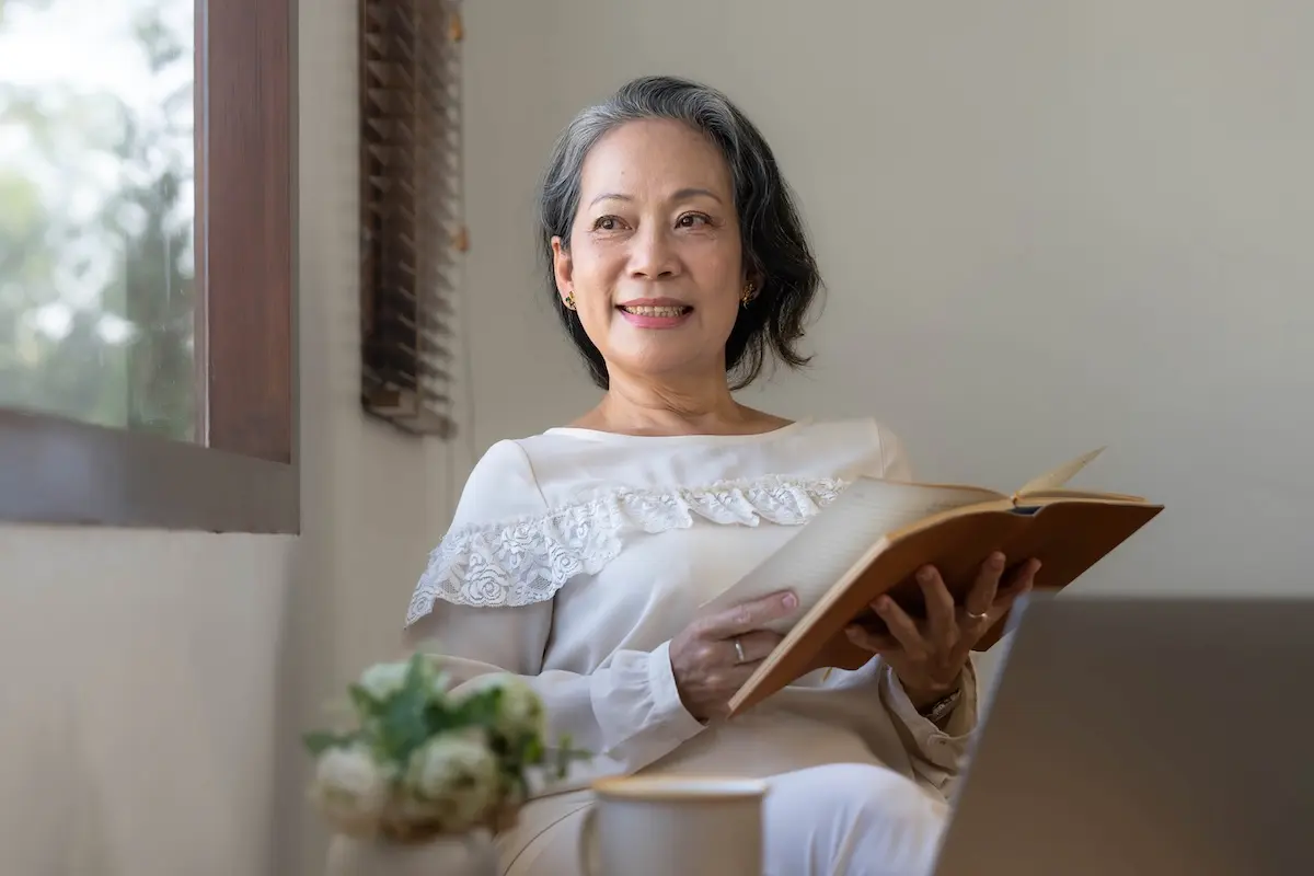 A smiling woman sitting by a window with a notebook, reviewing her financial plans and personal line of credit options.