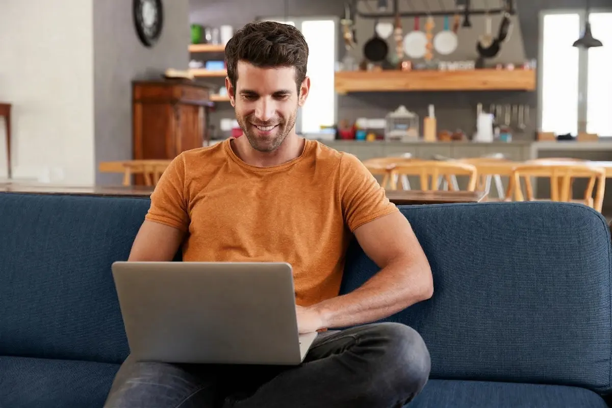 A smiling man sitting on a sofa and working on a laptop.