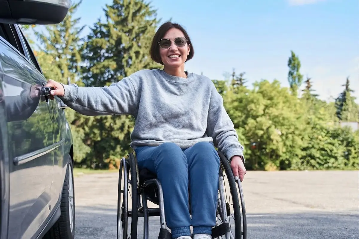 A smiling woman using a wheelchair opening a car door outdoors.