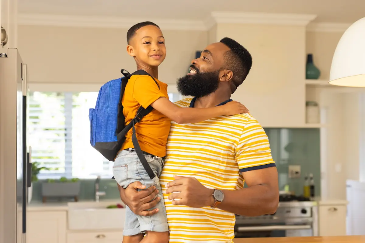 Father holding son with backpack, smiling together in the kitchen before school.