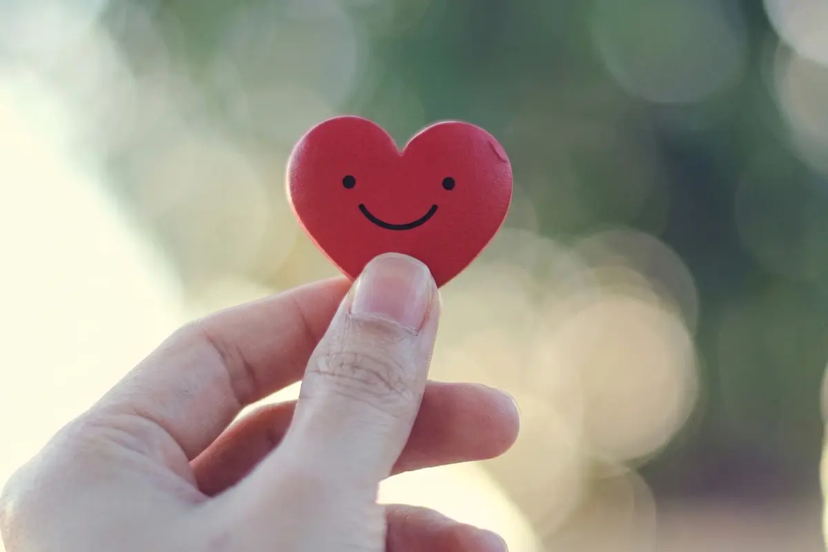 A hand is holding a small red heart-shaped object with a simple smiling face drawn on it.