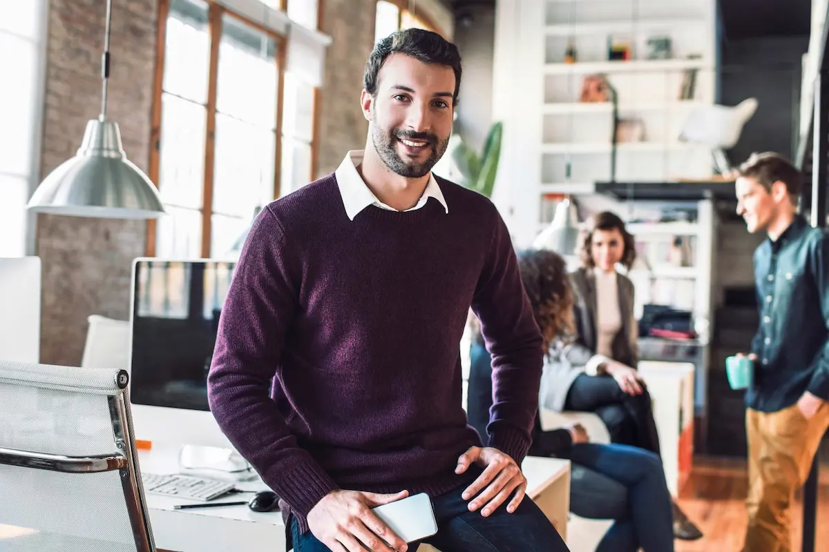 A man smiling and leaning on a desk in an office space.