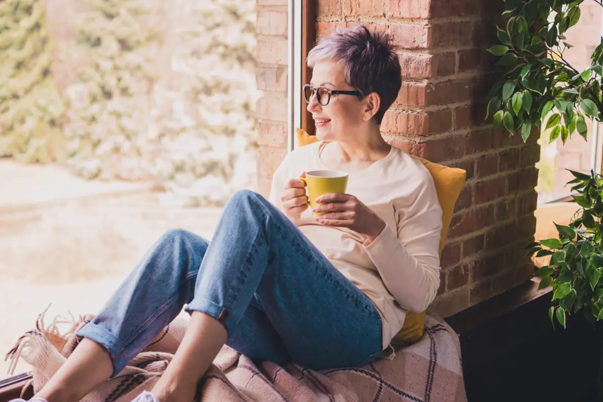 A woman sitting by a window holding a yellow coffee mug.