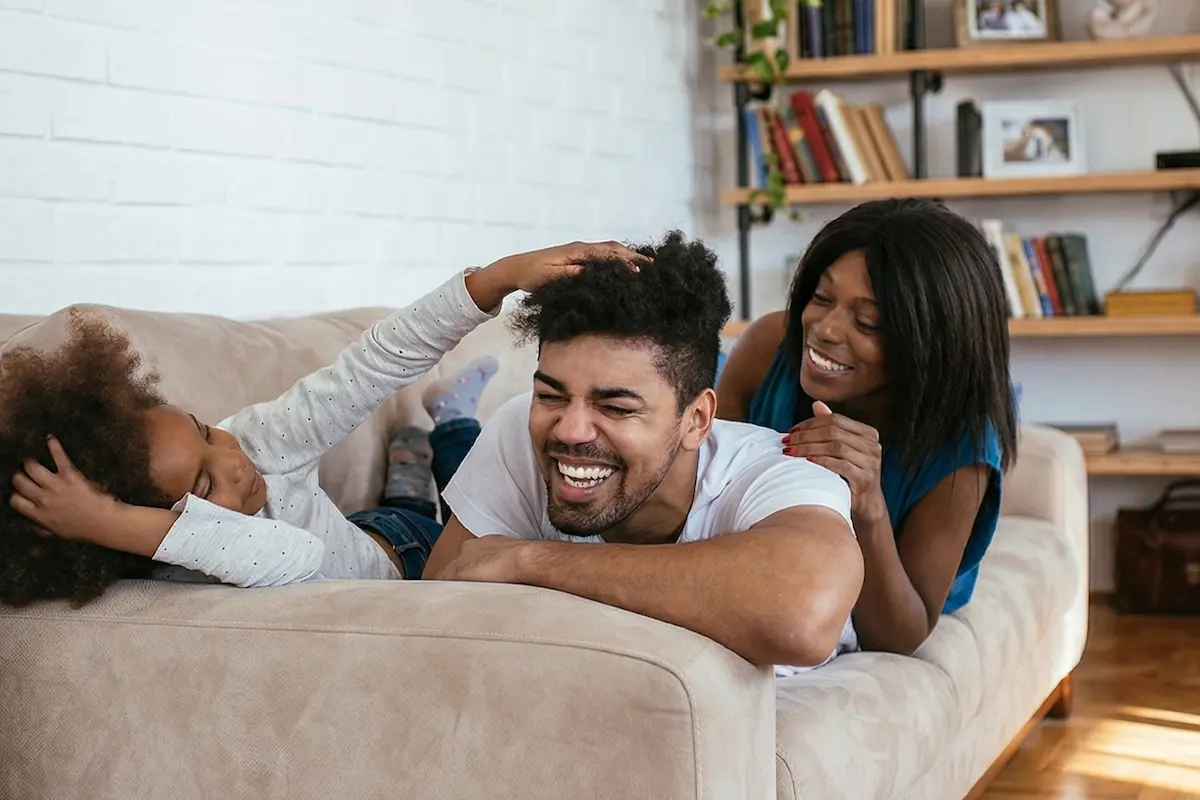 A joyful family of three playing and laughing together on a sofa in a living room.
