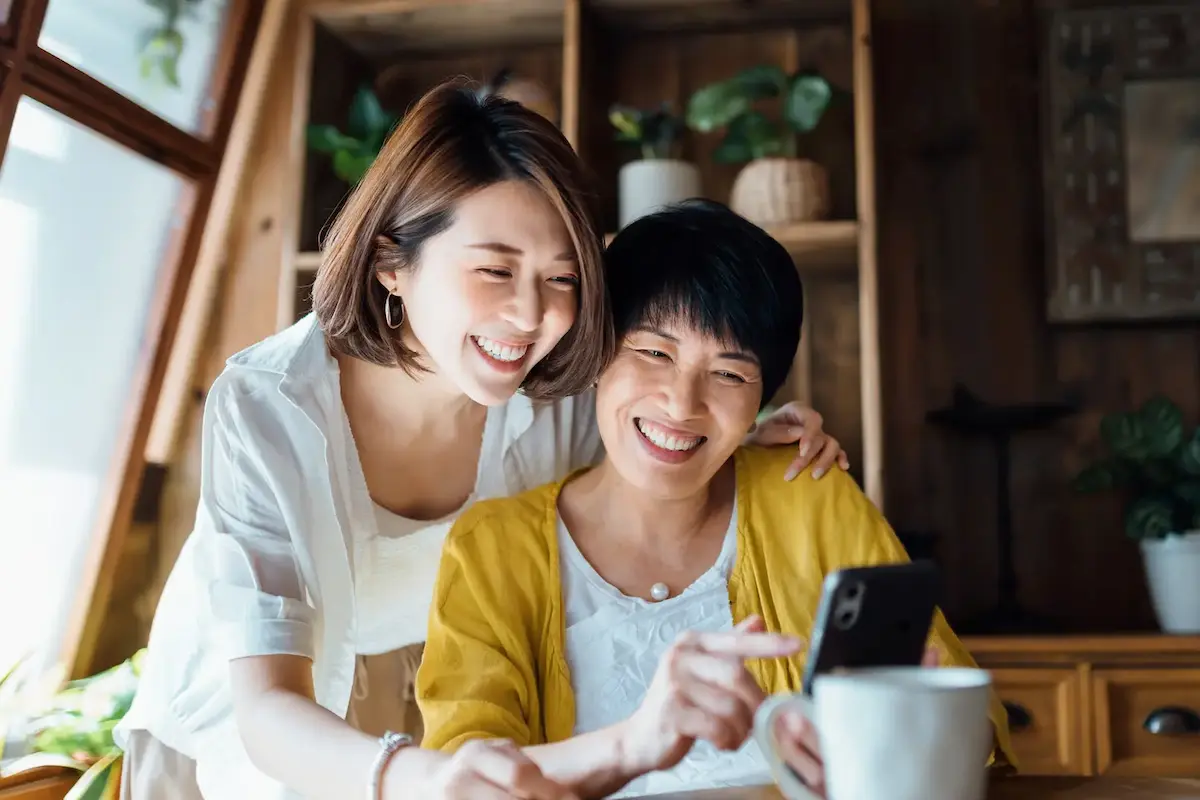 Two women at home smiling together while looking at a smartphone in a plant-filled room.