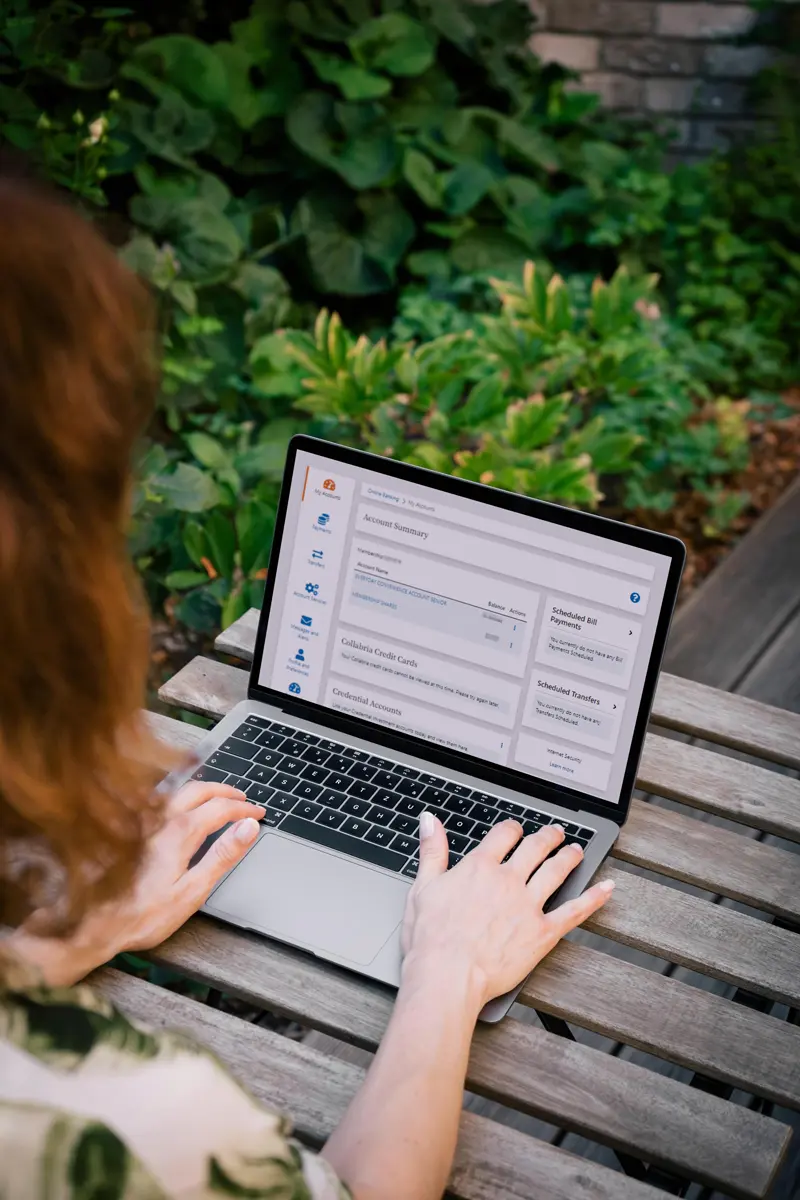 Person using a laptop on a wooden outdoor table, viewing the FirstOntario Credit Union online banking dashboard with account summary and transaction details, surrounded by green plants.