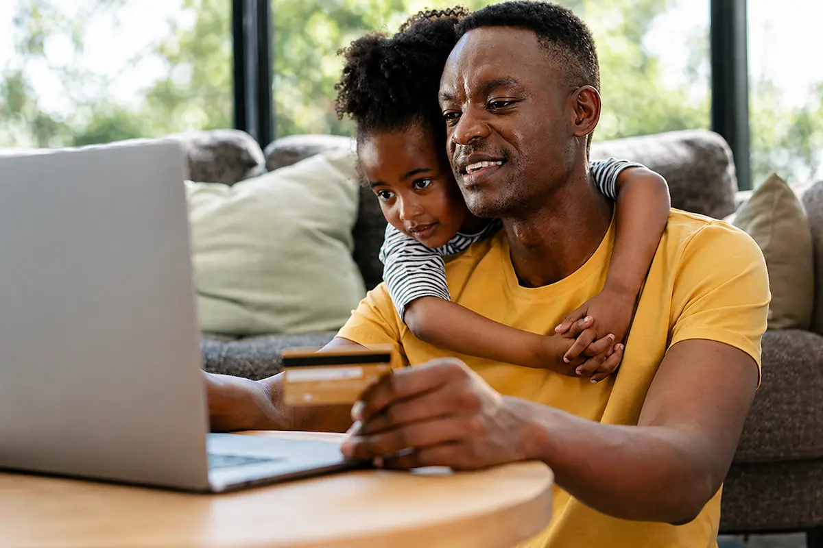 Father and young daughter using a laptop together while holding a credit card at home.